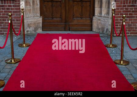 Sentiero con tappeti rossi con pietre dorate e corde di velluto rosso che conduce ad un grande ingresso storico in legno a doppia porta di un edificio in mattoni. Foto Stock
