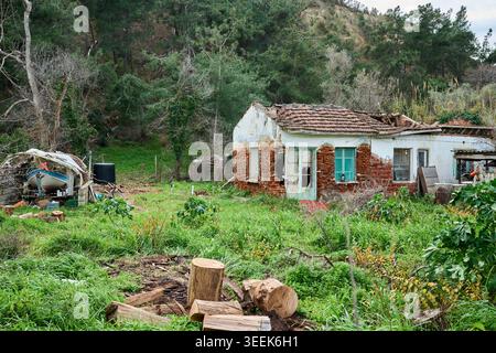 Una vecchia casa rurale abbandonata con un tetto piastrellato in rovina e mattoni esposti circondati da erba sovrastata e alberi verdi. Foto Stock