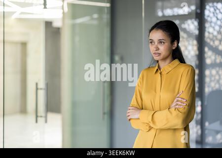 Giovane donna d'affari professionista in piedi con le braccia incrociate in un ufficio moderno, che guarda con attenzione mentre pianifica la crescita professionale, gli obiettivi di leadership e le ambizioni strategiche Foto Stock