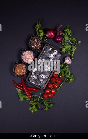 Pelmeni neri con impasto scuro e ripieno cremoso disposti in un vassoio tra pomodori ciliegini, cipolla rossa, aglio, prezzemolo, peperoncino e ciotole per spezie Foto Stock