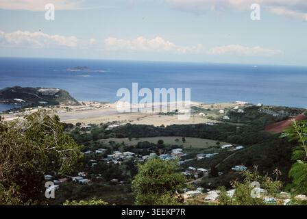 Una fotografia a colori d'archivio che si affaccia su un paesaggio costiero panoramico con una spiaggia, una baia, un piccolo insediamento costiero e colline sotto un cielo parzialmente nuvoloso. Foto Stock