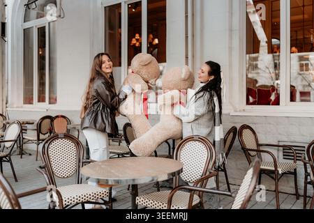 Friends Teddy Bear Cafe patio con due giovani donne sorridenti che tengono in mano giocattoli di peluche giganti durante una gioiosa gita invernale Foto Stock