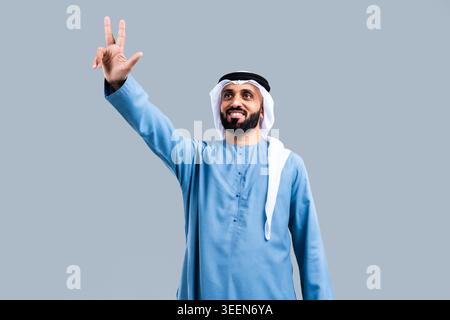 Uomo emirato barbuto che indossa kandura e ghutra tradizionali, sorridendo con sicurezza mentre alza una mano mostrando le dita per il segno V o vittoria, in una B. Foto Stock