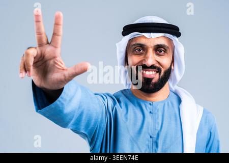 Sorridente uomo barbuto del Medio Oriente che indossa la tradizionale kandura e ghutra degli emirati, alzando la mano, formando l'unico gesto di vittoria d'amore usato Foto Stock