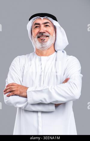Uomo anziano degli emirati sorridente guardando in alto con le braccia incrociate, ritraendo saggezza, successo e pensiero positivo per il futuro, isolato su uno sfondo grigio pulito Foto Stock