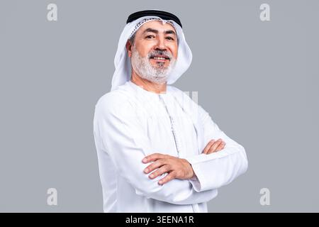 Uomo anziano degli emirati sorridente guardando la telecamera, indossando una kandora bianca e un ghutra, con le braccia incrociate su uno sfondo grigio chiaro, che rappresenta Foto Stock