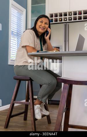 Donna asiatica matura che parla su smartphone mentre lavora all'isola della cucina con laptop e notebook Foto Stock