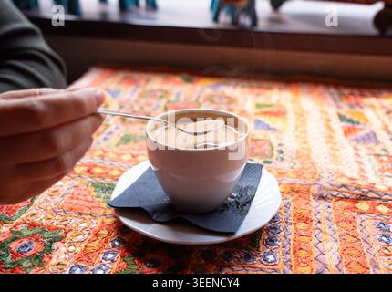 Una mano mescola tè o caffè caldo con il latte in una tazza bianca su un tavolo con un motivo tessile tradizionale su sfondo sfocato, messa a fuoco selettiva Foto Stock