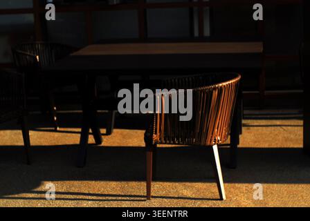 Una sedia in rattan e un tavolo in legno su un patio esterno, con una suggestiva luce del sole che crea ombre ad alto contrasto a terra. Foto Stock
