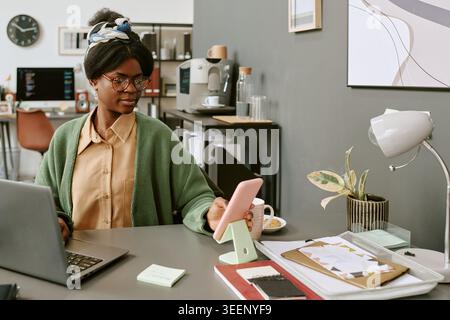 Donna adulta nera che controlla lo smartphone mentre lavora in ufficio Foto Stock