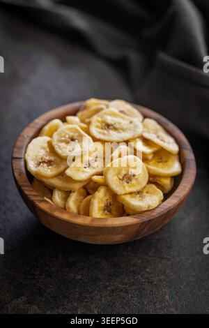 Patatine di banana croccanti ammucchiate in una ciotola di legno rustica su una superficie scura, che offre uno spuntino croccante e dolce alla frutta secca, perfetto per uno spuntino sano, pranzo Foto Stock