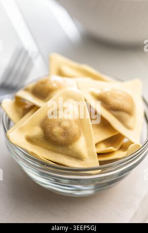 Ravioli freschi triangolari disposti su un tagliere di legno e in una ciotola di vetro, crudi e pronti da cucinare; l'impasto dorato evoca la comfor fatta in casa Foto Stock