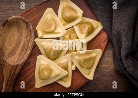Ravioli triangolari disposti su una tavola di legno con un cucchiaio di legno e un panno, evidenziando l'impasto fatto in casa crudo e i centri ripieni per rustici Foto Stock