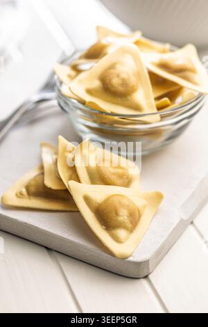 Ravioli freschi triangolari disposti su un tagliere di legno e in una ciotola di vetro, crudi e pronti da cucinare; l'impasto dorato evoca la comfor fatta in casa Foto Stock