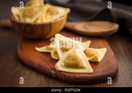 Ravioli triangolari disposti su una tavola di legno con un cucchiaio di legno e un panno, evidenziando l'impasto fatto in casa crudo e i centri ripieni per rustici Foto Stock