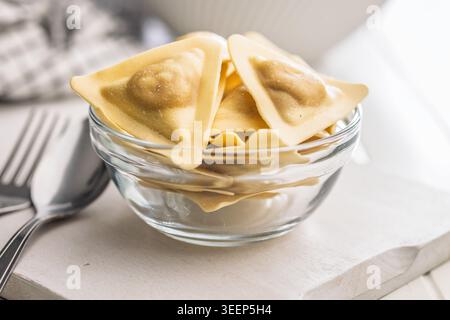 Ravioli freschi triangolari disposti su un tagliere di legno e in una ciotola di vetro, crudi e pronti da cucinare; l'impasto dorato evoca la comfor fatta in casa Foto Stock