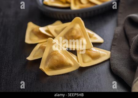Primo piano di ravioli triangolari su una superficie di legno scuro, evidenziando la pasta ripiena cruda con impasto dorato. Ideale per la cucina italiana, ricetta visua Foto Stock