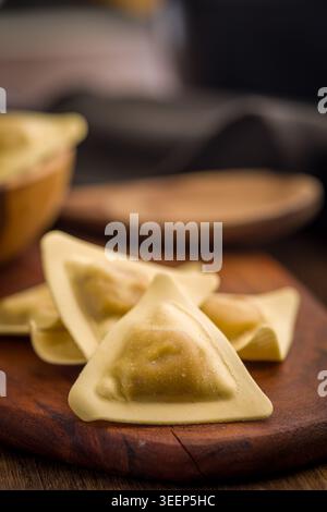 Ravioli triangolari disposti su una tavola di legno con un cucchiaio di legno e un panno, evidenziando l'impasto fatto in casa crudo e i centri ripieni per rustici Foto Stock
