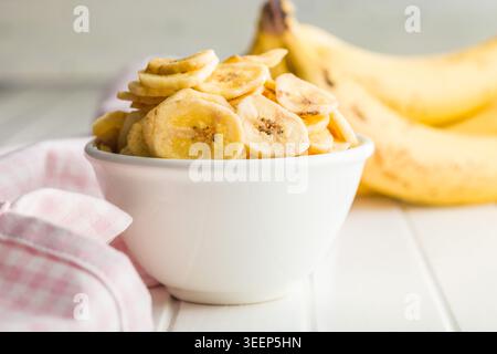 Una ciotola bianca piena di croccanti patatine di banana accanto a banane fresche mature e un panno rosa di Vichy su un tavolo bianco brillante, che evoca spuntini salutari, tro Foto Stock