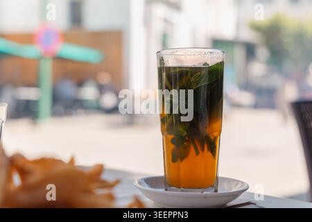 Rinfrescante tè alla menta marocchino con foglie fresche in un bicchiere alto, riposando su un piattino bianco in un caffè all'aperto Foto Stock
