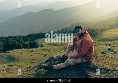 Un uomo barbuto che indossa un cappello e una giacca si siede su una collina erbosa durante l'ora d'oro al forte di Raghupur, Sojha, Manali, Himachal Pradesh, India. Foto Stock