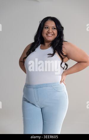 Donna sorridente e in posa in studio con canotta a coste bianca e pantaloni a vita alta azzurra Foto Stock