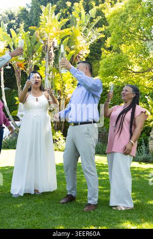 Sposa con gli ospiti del matrimonio che indossano abito e velo alzando flauti e brindando sul prato con champagne Foto Stock