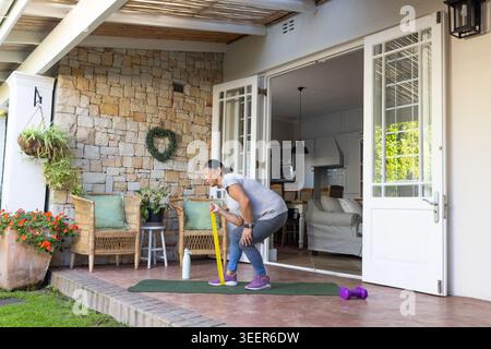 Donna afroamericana anziana che si esercita sul tappetino del patio tenendo asta, stupido e bottiglia in sportsw Foto Stock