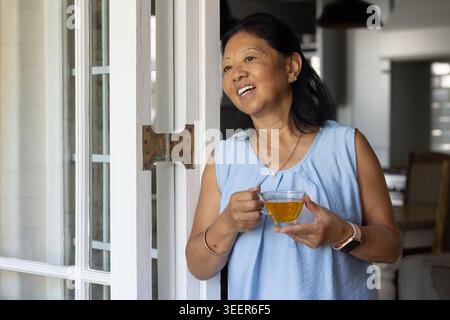 Donna asiatica anziana in piedi alla porta di vetro che tiene la tazza di vetro, che indossa il top azzurro con orologio Foto Stock