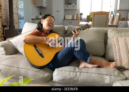 Donna asiatica anziana seduta su un divano grigio chiaro che suona la chitarra acustica e canta a casa Foto Stock