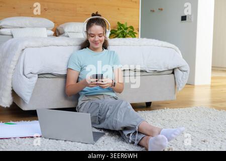 Donna adulta seduta su un tappeto in camera da letto, indossa le cuffie e utilizza smartphone e computer portatile Foto Stock
