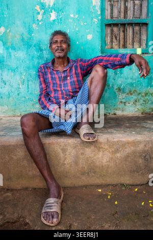 Uomo di mezza età che riposa fuori da una casa tradizionale dai colori vivaci in un villaggio rurale di Orissa, in India Foto Stock