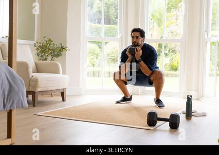 Uomo afroamericano che esegue squat su tappeto in soggiorno con un manichino esagonale indossando abbigliamento sportivo Foto Stock