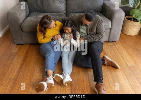 Famiglia di tre persone seduta su un pavimento di legno a casa, appoggiata verso il bambino vicino al divano grigio Foto Stock