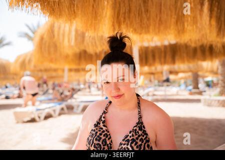 Donna caucasica in costume da bagno con stampa leopardo che posa su una spiaggia sabbiosa sotto ombrelloni di paglia in una località balneare. Turismo estivo e stile di vita per il tempo libero. Foto Stock