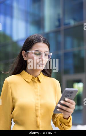 Giovane donna con una camicia gialla che indossa occhiali moderni e controlla il suo smartphone mentre cammina fuori da un ufficio, rappresentando la connettività urbana e la comunicazione digitale Foto Stock