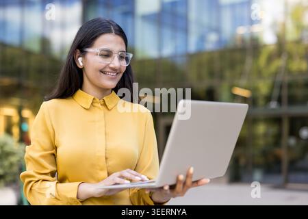 Giovane donna professionista che sorride, indossa occhiali e auricolari, lavora attivamente su un notebook all'esterno di un moderno edificio per uffici, rappresentando il lavoro a distanza e la tecnologia aziendale Foto Stock