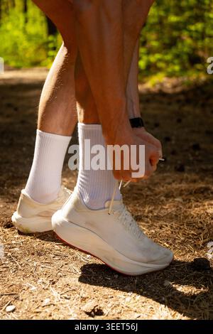 Primo piano di un corridore maschio che lega il pizzo della sua sneaker bianca a un sentiero soleggiato nella foresta. Concentrati su gambe muscolari, calze bianche e scarpe in setti boschivi Foto Stock