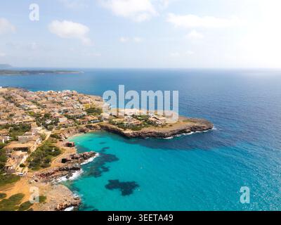 Vista aerea delle acque turchesi e della costa rocciosa della spiaggia con edifici residenziali e vegetazione lussureggiante sotto un cielo luminoso sa coma, Balear Foto Stock