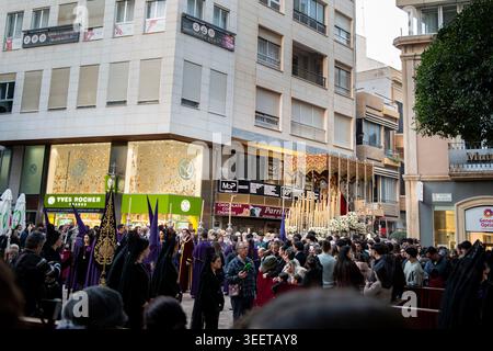 Almeria, Spagna - 30 marzo 2026: Le folle si riuniscono intorno a una processione della settimana Santa come nazarenos, penitenti e un galleggiante processionale con un'immagine sacra di Foto Stock