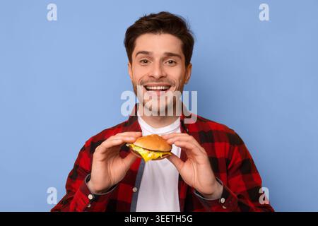L'uomo tiene Burger e sorrisi su sfondo blu mentre indossa una camicia a quadri rossa Foto Stock