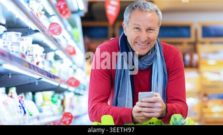 Senior Man Shopping nel negozio di alimentari mentre utilizza lo smartphone nel pomeriggio Foto Stock