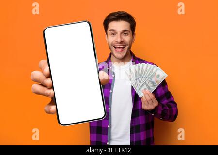 L'uomo tiene in mano denaro e smartphone mentre sorride su uno sfondo arancione in un ambiente luminoso Foto Stock