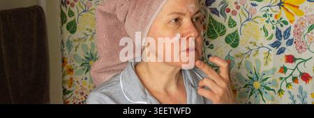 Una donna di 50 anni prepara la pelle applicando della crema nel suo bagno. Indossa un asciugamano sulla testa e si erge di fronte a un colorato curtai della doccia Foto Stock