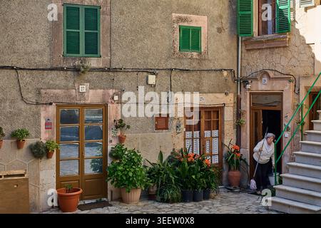 Maiorca, Spagna - 12 maggio 2026: Donna anziana che pulisce l'ingresso di una tradizionale casa in pietra mediterranea decorata con piante in vaso e verde Foto Stock