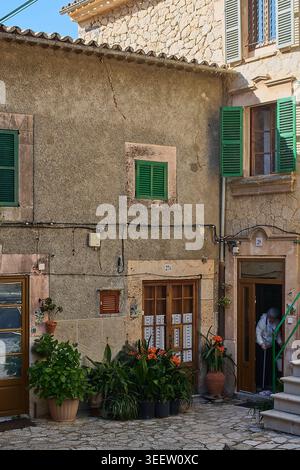 Maiorca, Spagna - 12 maggio 2026: Donna anziana che si imbatte all'interno della porta di una tradizionale casa in pietra mediterranea con persiane verdi e vasetti PL Foto Stock