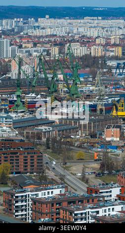 Vista aerea del porto industriale di Danzica con enormi gru da carico verdi, navi ancorate e un fitto paesaggio residenziale urbano con colline Foto Stock