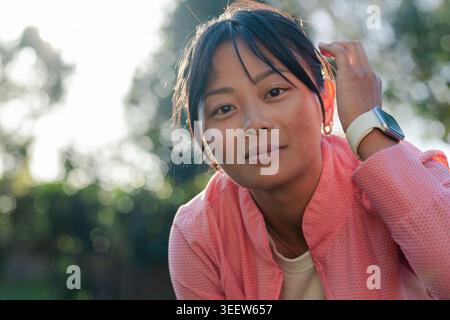 Donna asiatica adulta inclinata verso la macchina fotografica in giardino con giacca rosa corallo e smartwatch bianco Foto Stock