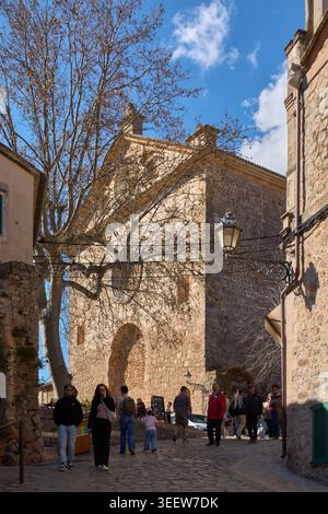 Maiorca, Spagna - 12 maggio 2026: I turisti camminano lungo una storica strada acciottolata verso la facciata in pietra di una chiesa tradizionale sotto un cielo blu Foto Stock
