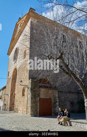 Maiorca, Spagna - 12 maggio 2026: Due turisti si siedono su una panchina di pietra di fronte alla storica chiesa di Sant Bartomeu, godendosi l'atmosfera tranquilla del Foto Stock